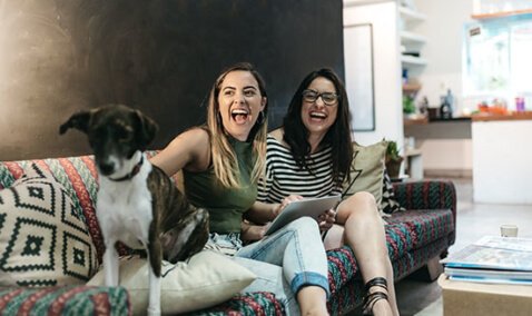 Two women laughing on a couch with a dog